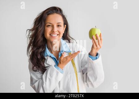 Lächelnde europäische Ärztin Ernährungswissenschaftlerin in weißem Mantel, die Apfel zeigt, über hellem Wandhintergrund steht und lächelt Stockfoto