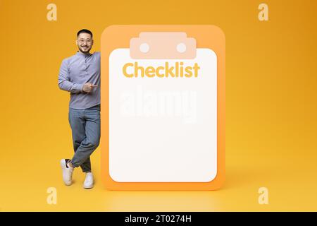 Ein lächelnder, erwachsener koreanischer Mann in Brille zeigt mit dem Finger auf eine große Checkliste mit Kopierraum Stockfoto