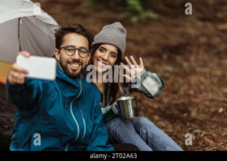 Positive junge kaukasische Familie in Jacken, die neben dem Zelt sitzt, Selfie am Telefon macht, Hand winkt Stockfoto
