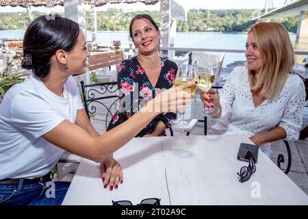 Drei fröhliche Freundinnen sitzen an einem Tisch mit einem Glas Wein, toasten, plaudern und lachen auf einer Restaurantterrasse am Fluss. Stockfoto