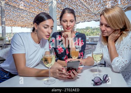 Drei fröhliche Freundinnen sitzen mit einem Glas Wein an einem Tisch und unterhalten sich auf einer Restaurantterrasse am Fluss. Bilder werden auf dem Telefon angezeigt. Stockfoto