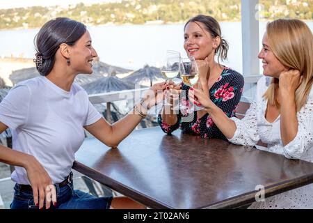 Drei fröhliche Freundinnen sitzen an einem Tisch mit einem Glas Wein, toasten, plaudern und lachen auf einer Restaurantterrasse am Fluss. Stockfoto