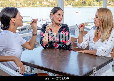 Drei fröhliche Freundinnen sitzen an einem Tisch mit einem Glas Wein, trinken, plaudern und lachen auf einer Restaurantterrasse am Fluss. Stockfoto