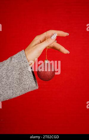 Rote runde Weihnachtsbaumkugel in weiblicher Hand isoliert auf rotem Hintergrund. Die Hand einer Frau hält ein dekoratives Weihnachtsbaumspielzeug. Neujahr und Weihnachten Stockfoto