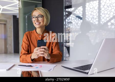 Junge schöne Frau am Arbeitsplatz, die Anwendung am Telefon verwendet, Geschäftsfrau träumerisch denkt, die aus dem Fenster blickt, Smartphone hält, im Büro mit Laptop arbeitet. Stockfoto