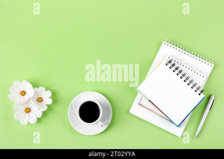 Künstlerische Komposition mit offenem leerem Notizblock und Stift, Tasse schwarzen Kaffee, Blumen eines weißen Kosmos auf grünem Hintergrund. Festlicher Bürotisch c Stockfoto