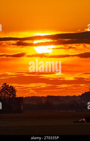 Wunderschöner wolkig-roter Sonnenuntergang mit gigantischer Sonne über großen Feldern und Bäumen, gesehen von einem 600-mm-Kameraobjektiv Stockfoto