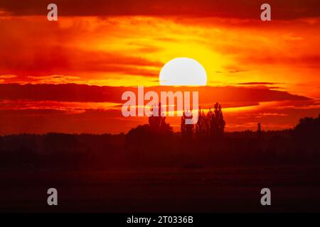 Wunderschöner wolkig-roter Sonnenuntergang mit gigantischer Sonne über großen Feldern und Bäumen, gesehen von einem 600-mm-Kameraobjektiv Stockfoto