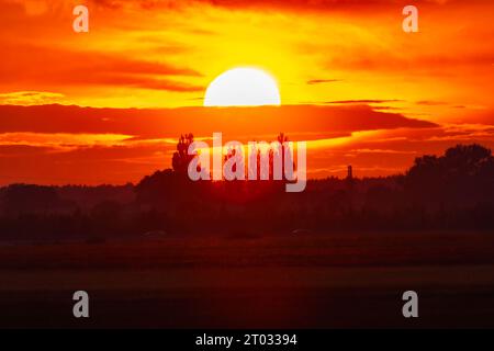 Wunderschöner wolkig-roter Sonnenuntergang mit gigantischer Sonne über großen Feldern und Bäumen, gesehen von einem 600-mm-Kameraobjektiv Stockfoto