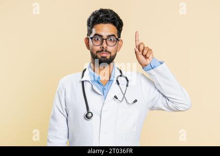 Eureka. Inspirierter junger indischer Arzt Kardiologe Mann, der mit offenem Mund den Finger nach oben zeigt, gute Idee, Plan, Start-up, Inspiration Motivation Lösung hat. Apotheke-Typ auf beigefarbenem Hintergrund Stockfoto