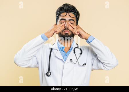 Erschöpft müde indische junge Doktor Kardiologe Mann zieht die Brille aus, fühlt Augenschmerzen, ist Überarbeit Burnout von langer Arbeit. Schläfriger, erschöpfter Kerl, der die Augen isoliert auf beigefarbenem Hintergrund reibt Stockfoto