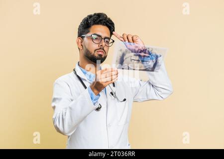 Der indische junge Arzt Kieferorthopäden untersucht ein Panorama-Röntgenbild der Kieferzähne. 3D-Modell des Mundes des Patienten, MRT-Scan. Zahnheilkunde, Mundpflege. Ein arabischer Stomatologe auf beigefarbenem Hintergrund Stockfoto