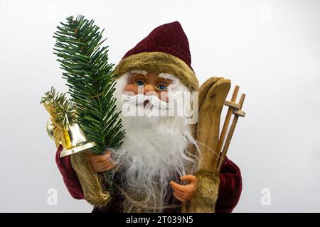 Das Weihnachtsmann-Spielzeug hält Holzskier in der einen Hand und einen Tannenzweig mit einer Glocke in der anderen auf weißem Hintergrund Stockfoto