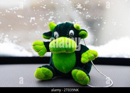 Grüne Spielzeugkuh, die auf dem Armaturenbrett eines Autos sitzt, während draußen schneit Stockfoto