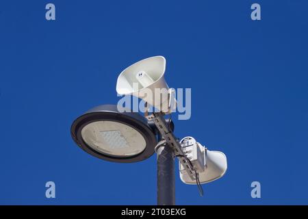 Detail des oberen Teils einer Straßenlaterne, die ein Paar Lautsprecher unterstützt, um die Bevölkerung über den blauen Himmel im Hintergrund zu informieren Stockfoto