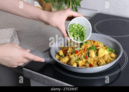 Frau fügt geschnittene grüne Zwiebeln zu Reis mit Fleisch und Gemüse in der Pfanne hinzu, Nahaufnahme Stockfoto