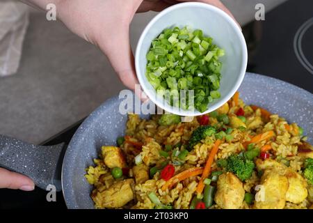 Frau fügt geschnittene grüne Zwiebeln zu Reis mit Fleisch und Gemüse in der Pfanne hinzu, Nahaufnahme Stockfoto