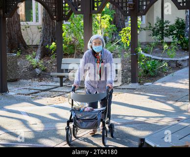 Seniorin mit Gesichtsmaske, die mit ihrem Gehhilfen in einem Pensionsdorf draußen läuft. Stockfoto