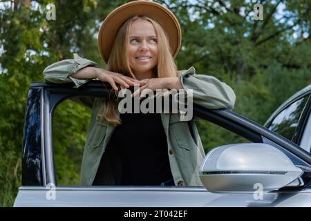 Blonde Frau mit Hut, die neben der Autotür wohnt. Junge Touristen erkunden lokale Reisen und machen echte Momente. Wahre Emotionen Ausdruck des Ausgehens und der Erfrischung Entspannen Sie sich an der frischen Luft Stockfoto