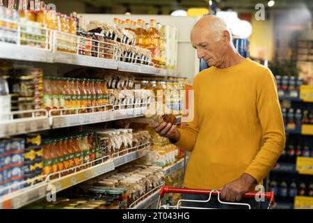 Älterer Mann im Supermarkt kaufte Lebensmittel mit einem Glas Salat, der im Supermarkt am Gang entlang ging Stockfoto