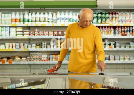 Lebensmittelkauf-Konzept. Älterer Mann, der im Supermarkt einkaufen, im Supermarkt kaufen, wählen Sie die Produkte, die in der Nähe des Kühlschranks im Hypermarkt stehen Stockfoto