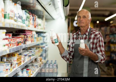 Porträt eines lächelnden alten Arbeiters auf Schürze, der Milchprodukte im Supermarkt hält Stockfoto