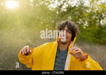 Positiver junger Mann lächelt in gelber Regenjacke während des Regens im Park. Genießen Sie den Regen im Freien. Der Araber fängt die Regentropfen mit Stockfoto