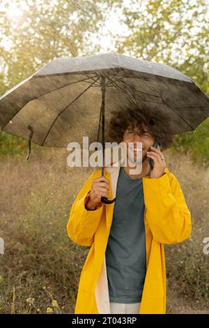 Ein arabischer Mann in einem gelben Regenmantel unter einem Regenschirm, der auf einem Handy spricht und lächelt Stockfoto