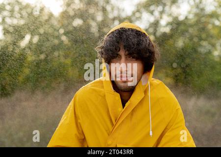 Trauriger Mann friert im Regen mit gelbem Regenmantel im Park ein. Gutaussehender Mann unter Regen draußen. Der Typ hat einen müden Ausdruck während des Regens Stockfoto
