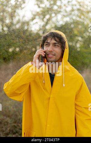 Ein arabischer Mann in einem gelben Regenmantel im Regen, der auf einem Handy spricht, kommuniziert mit einem Freund Stockfoto