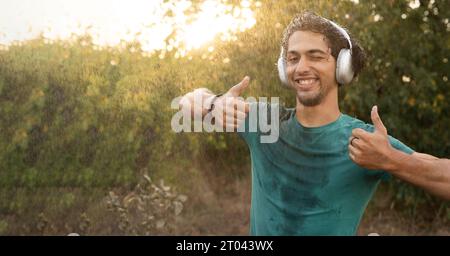 Porträt eines fröhlich lächelnden jungen arabischen Mannes, der Musik im Regen genießt und Daumen mit Klassengesten zeigt Stockfoto