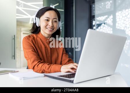 Junge erfolgreiche Programmiererin, die im Büro mit Laptop arbeitet, asiatische Frau im Kopfhörer hört Online-Musik und Hörbücher, arbeitet im Büro, lächelt zufrieden. Stockfoto