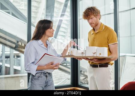 Zwei kreative Teammitglieder in eleganten Outfits arbeiten an einem maßstabsgetreuen Modell des Gebäudes, Designbüro Stockfoto