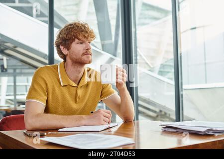 Nachdenklicher junger Mann mit roten Haaren, der an seinen Dokumenten arbeitet und eine Teetasse hält, Coworking-Konzept Stockfoto