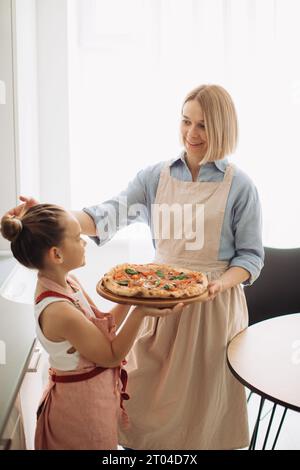 Eine schöne Mutter und ihre süße Tochter halten eine frisch gebackene Pizza. Konzept des Kochens zu Hause, Stockfoto