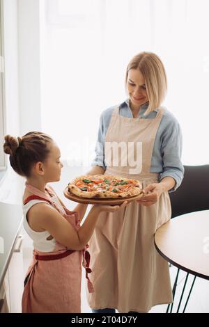 Eine schöne Mutter und ihre süße Tochter halten eine frisch gebackene Pizza. Konzept des Kochens zu Hause, Stockfoto