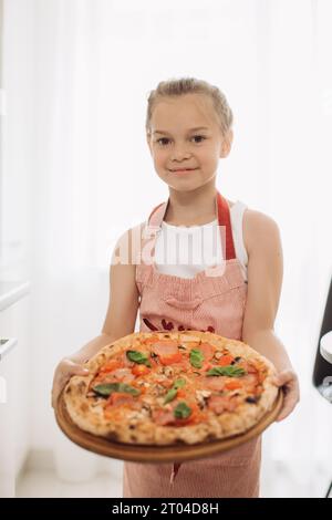 Eine kleine 8-jährige Köchin hält eine köstliche Pizza, während sie in einer modernen, hellen Küche steht. Stockfoto