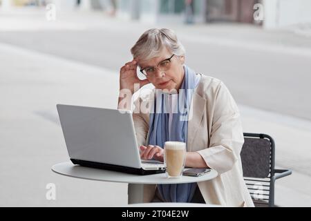 Eine nachdenkliche Geschäftsfrau mit grauen kurzen Haaren arbeitet am Laptop, schaut in den Computer am Tisch im Café im Freien. Alter in der Technik. Stockfoto