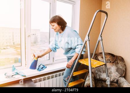 Glückliche, Schüchterne Frau, Die Fenster Mit Spray Reinigt. Active Seniors-Konzept. Reinigungsappartement Für Ältere Frauen. Hausfrau. Kaukasische Frau Im Mittleren Alter Stockfoto