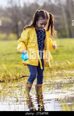 Ruhiges Kind, in gelber Jacke und Gummistiefeln, stehend in einer Pfütze mit blauem Papierboot in der Hand und schaute auf Wasser, bereitete sich vor, Shi zu starten Stockfoto