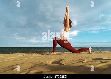 Eine junge Frau in ihren Zwanzigern spielt die Yoga-Pose Sun Salutation am Sandstrand. Profilaufnahme fängt das ruhige Meer, den Horizont und die farbenfrohe SK ein Stockfoto