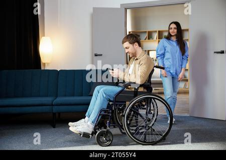 Ein süßer junger Mann im Rollstuhl hält ein Handy, während seine Freundin ihm hilft, sich in der Wohnung zu bewegen. Stockfoto