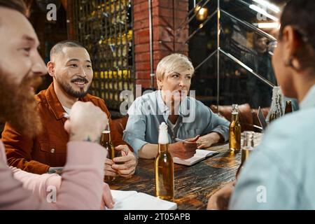 Kreatives multikulturelles Team, das in einem Pub in der Nähe von Bierflaschen sitzt und neues Geschäftsprojekt diskutiert Stockfoto