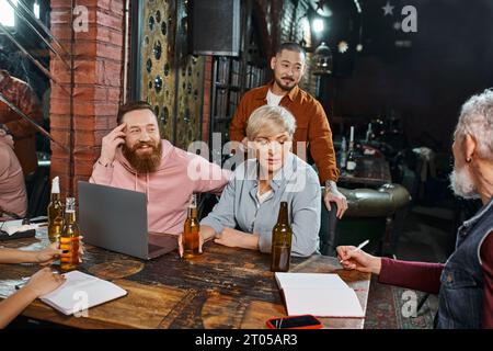 Multikulturelles Team, das in einem Pub neben Bierflaschen und Laptop sitzt und neues Geschäftsprojekt diskutiert Stockfoto