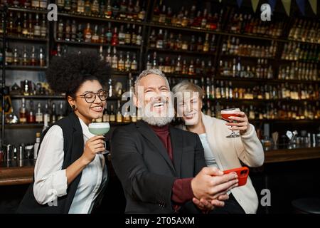 Fröhlicher bärtiger Mann, der Fotos mit multiethnischen Frauen in der Bar macht, ein vielfältiges Team, das sich nach der Arbeit ausruhen lässt Stockfoto