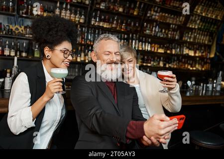 Fröhliche multiethnische Frauen mit Cocktails in der Nähe eines bärtigen Kollegen, die Selfie auf dem Smartphone in der Bar machen Stockfoto