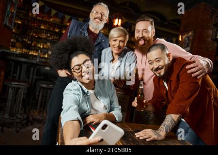 Lustige afroamerikanische Frau, die die Zunge ausstreckt und Selfie mit multiethnischen Arbeitskollegen in der Bar macht Stockfoto