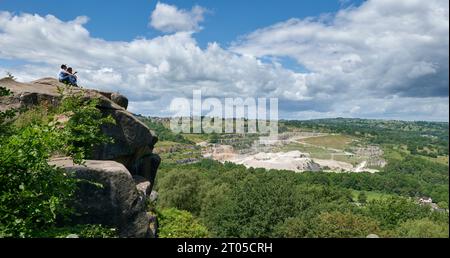 Besucher, die auf Black Rocks in der Nähe von Cromford, Derbyshire, sitzen, wo Kalkstein aus dem Dene Quarry in der Ferne abgebaut wird Stockfoto