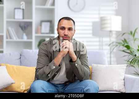 Porträt eines jungen hispanischen Mannes, der ernsthaft in die Kamera schaut, zu Hause mit gefalteten Händen auf der Couch sitzt. Stockfoto