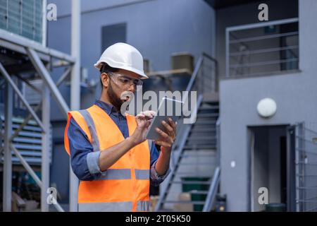 Ernsthafter denkender und fokussierter Ingenieur, der in der Fabrik mit Schutzhelm und Weste arbeitet, Mann mit Tablet-Computer, der Diagramme liest, und Ausrüstung in der industriellen Fabrik überprüft. Stockfoto